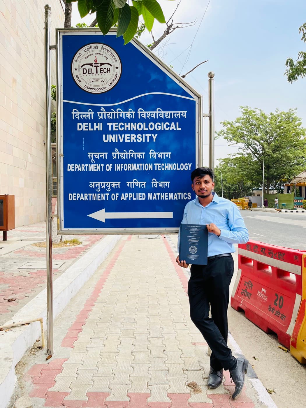Aftab holding his degree beside the Delhi Technological University sign.